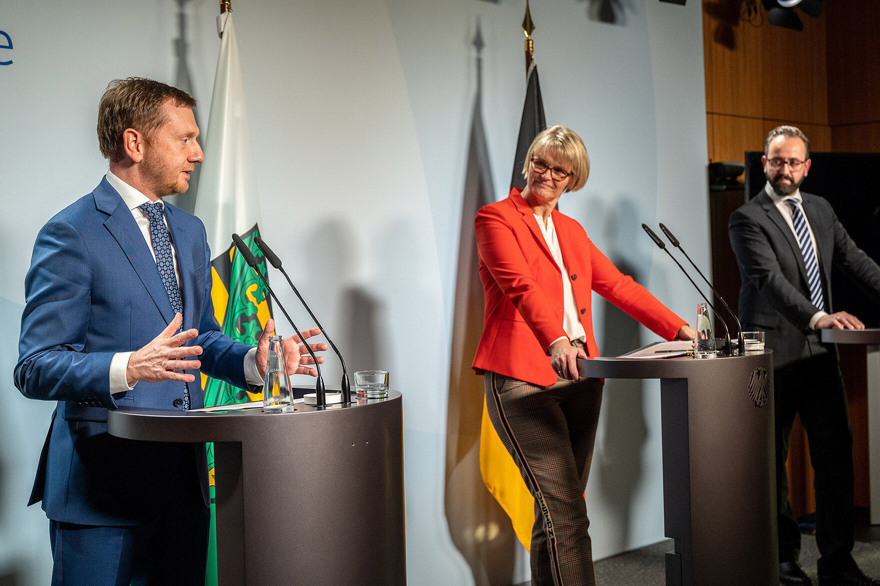 Zwei Männer und eine Frau stehen an Rednerpulten. Einer der Männer spricht und gestikuliert. Im Hintergrund stehen Fahnen des Freistaates Sachsen und der Bundesrepublik Deutschland.